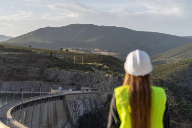 Güvenlik yeleği ve kask giyen kadın mühendis, dağlık bir arazide barajı inceliyor, yapısal bütünlüğü ve su yönetimini sağlıyor.