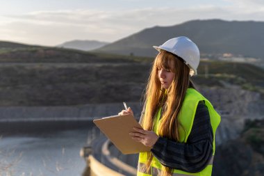 Güvenlik yeleği ve kask giyen genç bir kadın mühendis barajı inceliyor, bir panoya not alıyor, yapısal bütünlüğü ve su kaynakları yönetimini sağlama alıyor.