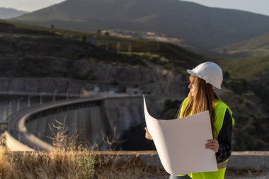 Güvenlik yeleği ve kask giyen kadın mühendis hidroelektrik barajının yakınındaki planı inceliyor, altyapı ve yenilenebilir enerji geliştirme üzerine odaklanıyor