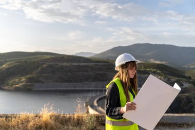 Güvenlik yeleği ve kask giyen kadın mühendis, bir baraj barajının yakınındaki planları inceleyerek, yapısal bütünlüğü ve operasyonel verimliliği garanti altına alıyor.