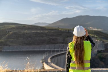 Güvenlik yeleği giyen kadın mühendis, dağlık bir arazide barajı teftiş ederek, yapısal bütünlüğü ve su kaynakları yönetimini sağlama alıyor.