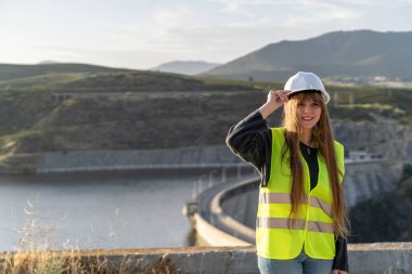 Güvenlik yeleği ve şapka takan genç bir kadın mühendis, güvenli bir şekilde bir baraj ve sakin bir gölün yanında duruyor, altyapı alanında profesyonelliği somutlaştırıyor.