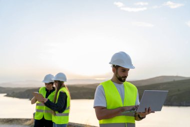 Günbatımında göl kenarında dizüstü bilgisayar ve tablet kullanan mühendisler, mühendislik ve teknoloji alanlarındaki kariyer fırsatlarını vurguluyorlar.