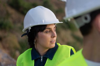 Güvenlik yeleği ve şapka giyen kadın mühendis inşaat alanındaki meslektaşımla proje detaylarını tartışıyor.