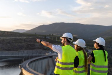 Güvenlik yelekleri ve kask giyen üç mühendis gün batımında dağlardaki bir hidroelektrik santralini kontrol ediyor.