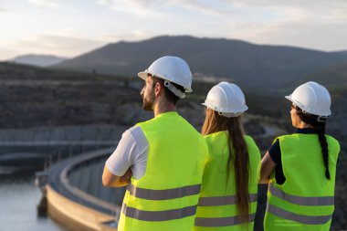 Çelik yelek giyen mühendisler bir dağ vadisinde baraj ve su deposunu teftiş ediyorlar.