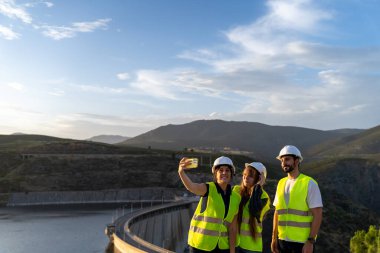 Güvenlik yelekleri ve kasklar takan mühendisler, arka planda dağlar olan bir baraj inşaatında selfie çekiyorlar.
