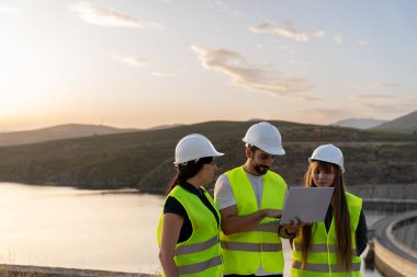 Güvenlik yeleği ve kask giyen üç mühendis gün batımında hidroelektrik santrali barajının yanında dizüstü bilgisayar kullanarak bir projeyi tartışıyorlar.