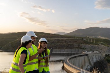Güvenlik yelekleri ve kasklar takan mühendisler dizüstü bilgisayar kullanıyor, gün batımında hidroelektrik santrali barajının yakınındaki verileri analiz ediyorlar.