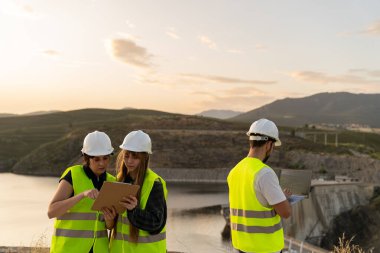 Gün batımında bir inşaat sahasında dijital cihazlar kullanarak güvenlik yeleği ve kask takan mühendisler, inşaat mühendisliğinde teknoloji entegrasyonunu gösteriyor.