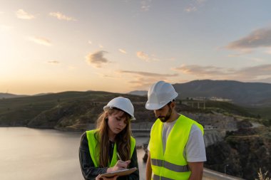 Güvenlik yeleği ve kask giyen iki mühendis, veri toplama ve analiz için dijital tablet ve pano kullanarak gün batımında bir su deposu barajını inceliyor.