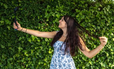 Yeşil sarmaşık duvarının önünde kamera ve selfie çubuğu taşıyan genç bir kadın takipçileri için yaz içeriği yaratıyor.