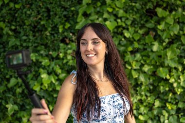 Genç gülümseyen etkili yeşil sarmaşık duvarının önünde selfie çubuğuyla online içerik yaratıyor.