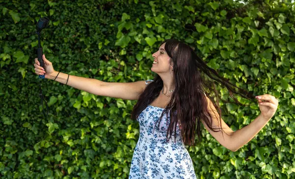 Yeşil sarmaşık duvarının önünde kamera ve selfie çubuğu taşıyan genç bir kadın takipçileri için yaz içeriği yaratıyor.
