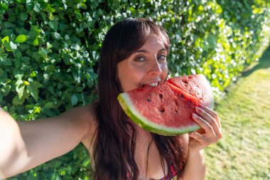 Güneşli bir yaz gününde bir dilim taze karpuzun tadını çıkaran genç bir kadın, bir selfie ile anı yakalıyor.
