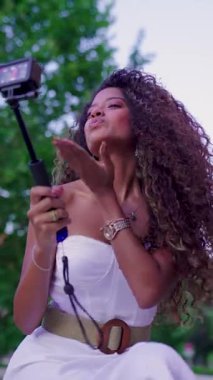 Uzun kıvırcık saçlı ve beyaz elbiseli nüfuzlu. Madrid 'de bir parkta selfie çubuğuyla içerik yaratıyor.