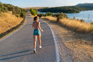 Formda bir kadın güneşli bir yaz gününde açık havada koşuyor, manzaranın tadını çıkarıyor ve çevresindeki doğanın tadını çıkarıyor.