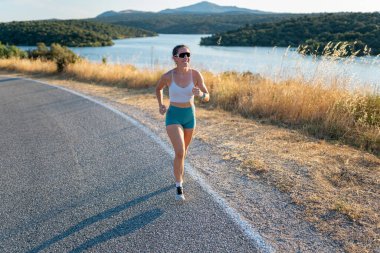 Genç bir kadın güneşli bir yaz gününde asfalt yolda koşuyor, manzaranın tadını çıkarıyor.