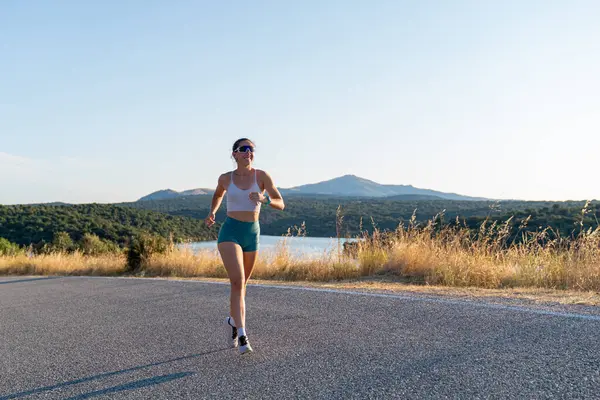Formda bir kadın göl kenarında manzaralı bir yolda koşuyor, doğa ile çevrili bir yaz akşamı egzersizinin tadını çıkarıyor.