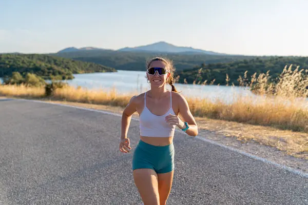 Güneşli bir günde dışarıda koşan zinde bir kadın, yuvarlanan tepelerle çevrili sakin bir gölün güzel manzarasından zevk alıyor.