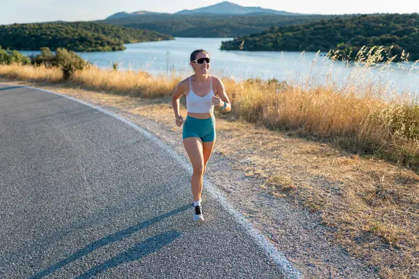 Genç bir kadın güneşli bir yaz gününde asfalt yolda koşuyor, manzaranın tadını çıkarıyor.