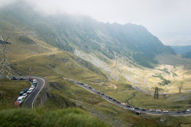 Dönek bir dağ yolunda, bulutlu bir günde, yemyeşil manzara ve sisle çevrili arabalar sürüp kuyruğa giriyor.