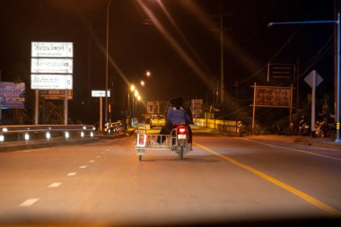 Sidecar 'ı olan bir motosiklet gece gökyüzünün altında sokak lambalarıyla aydınlatılmış boş bir yolda ilerler. Arka plandaki işaretler kırsal bölgeleri ve yerel yönleri gösteriyor..