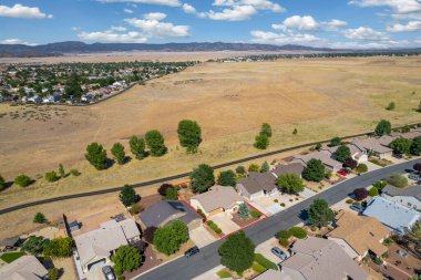 Geniş açıdan bakıldığında, ağaçların arasına yerleştirilmiş birkaç evi olan sakin bir yerleşim yeri görülüyor. Gündüz vakti, açık mavi gökyüzünün altındaki açık arazi ve tepeleri de kapsıyor..