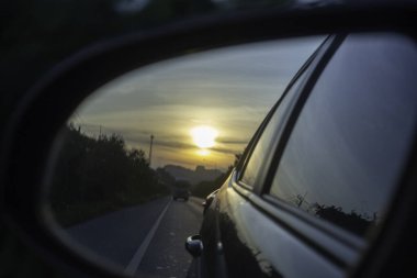 Huzurlu bir akşam gezintisi arabaların sakin bir yoldan geçerken canlı renkleri yansıtan, yan aynaya yansıtılan güzel bir günbatımı sergiliyor..