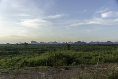 Berrak mavi bir gökyüzünün altında yemyeşil alanlar yayıldı, engebeli dağların silueti ufukta belirdi..