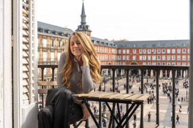 Bir kadın zarif bir şekilde rahat bir balkonda oturuyor, önünde uzanan hareketli şehrin güzel manzarasının tadını çıkarıyor.