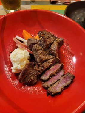 Bir masada, üzerinde zarif bir kırmızı tabak ve yanında kremalı patates püresi ile birlikte lezzetli bir biftek var.