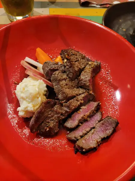 Bir masada, üzerinde zarif bir kırmızı tabak ve yanında kremalı patates püresi ile birlikte lezzetli bir biftek var.