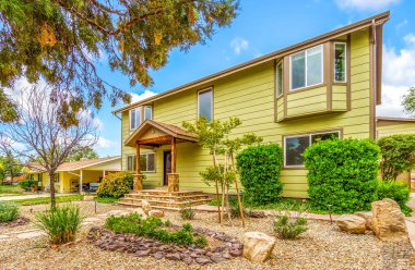 Geniş, büyük sarı bir ev. Önünde çok sayıda pencere ve yemyeşil çalılar var.