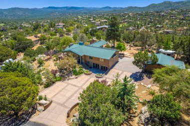 Geniş bir hava manzarası, bereketli ağaçların ve görkemli dağların arasına yerleşmiş sakin bir ev sergiliyor.