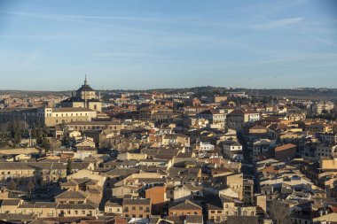 Geniş bir hava manzarası, geniş mavi gökyüzü altında binalar ve çatılarla dolu kentsel bir manzara sunan canlı bir şehir gösteriyor.