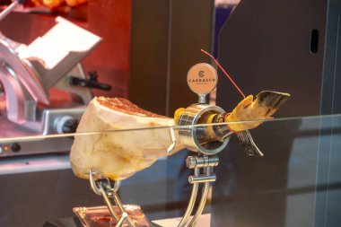 Lezzetli bir şekilde hazırlanmış bir parça jambon, bir et dilimleme makinesinin tam önünde duran cam tezgahın üzerinde zarif bir şekilde dinleniyor.