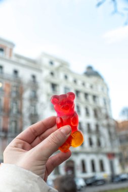 Bir insan, büyük bir binanın önünde parlak kırmızı bir jelibonu elinde tutuyor ve izleyicilere lezzetli şekerleri gösteriyor.