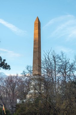 Yükselen, büyük bir dikilitaş, berrak mavi gökyüzünün altında görkemli yeşil ağaçlarla çevrili, resim gibi bir manzara yaratıyor.