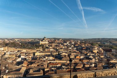 Geniş bir hava manzarası, geniş mavi gökyüzü altında binalar ve çatılarla dolu kentsel bir manzara sunan canlı bir şehir gösteriyor.