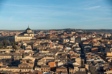Geniş bir hava manzarası, geniş mavi gökyüzü altında binalar ve çatılarla dolu kentsel bir manzara sunan canlı bir şehir gösteriyor.