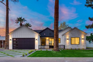 Zarif, siyah bir garaj kapısı olan büyüleyici beyaz bir ev güzel bir şekilde bereketli palmiye ağaçları ile çevrili ve güzel bir manzara yaratıyor.