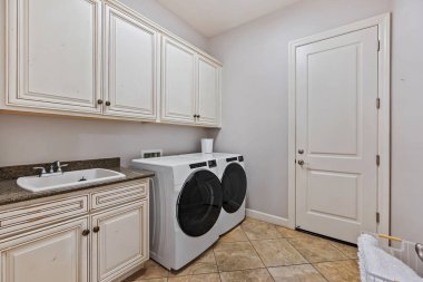 Bu çamaşır odasında bolca dolap, yıkanacak uygun bir lavabo ve içeri ışık girmesini sağlayan parlak bir pencere var.