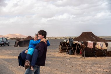 Bir adam, kollarında bir çocuğu nazikçe taşır onlar güneşin altında, uçsuz bucaksız ve çorak çöl arazisinde yol alırken.