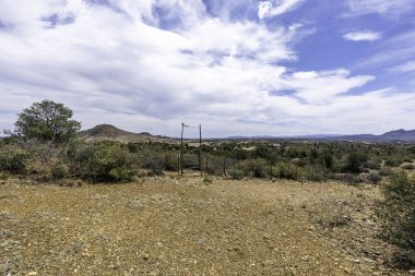 Uçsuz bucaksız bir çöl manzarası etkileyici dağları barındırır. Arkaplanda, kabarık bulutlarla süslenmiş canlı mavi bir gökyüzünün altında.