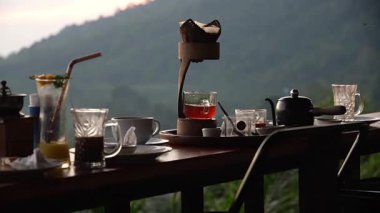 Videoda güzel düzenlenmiş bir masası, kahve makinesi, fincanları ve dingin bir dağ manzarasına karşı hazırlanmış sevimli bir çaydanlığı olan rahat bir kafe yer alıyor.