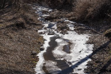 Bir su akıntısı donmuş ve çamurlu bir görünüme sahip. Su tepeden aşağı akıyor ve etrafı kayalarla çevrili.
