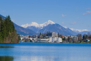 Sol arka plandaki ağaçların arasında gölü ve karlı dağları olan kanlı bir şehir.
