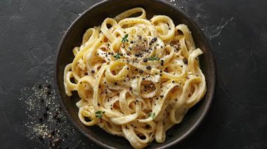 Kremalı makarna tabağının yakın plan çekimi, muhtemelen fettuccine alfredo, karabiber ve kekik ile süslenmiş, koyu bir arka planda koyu bir kasede servis ediliyor..