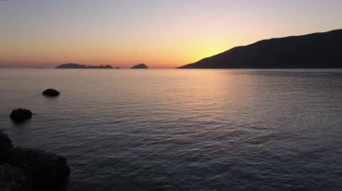 Sakin bir denizin üzerinde sakin bir güneş doğuyor. Uzak adalar ve dağların silueti canlı gökyüzüne karşı..
